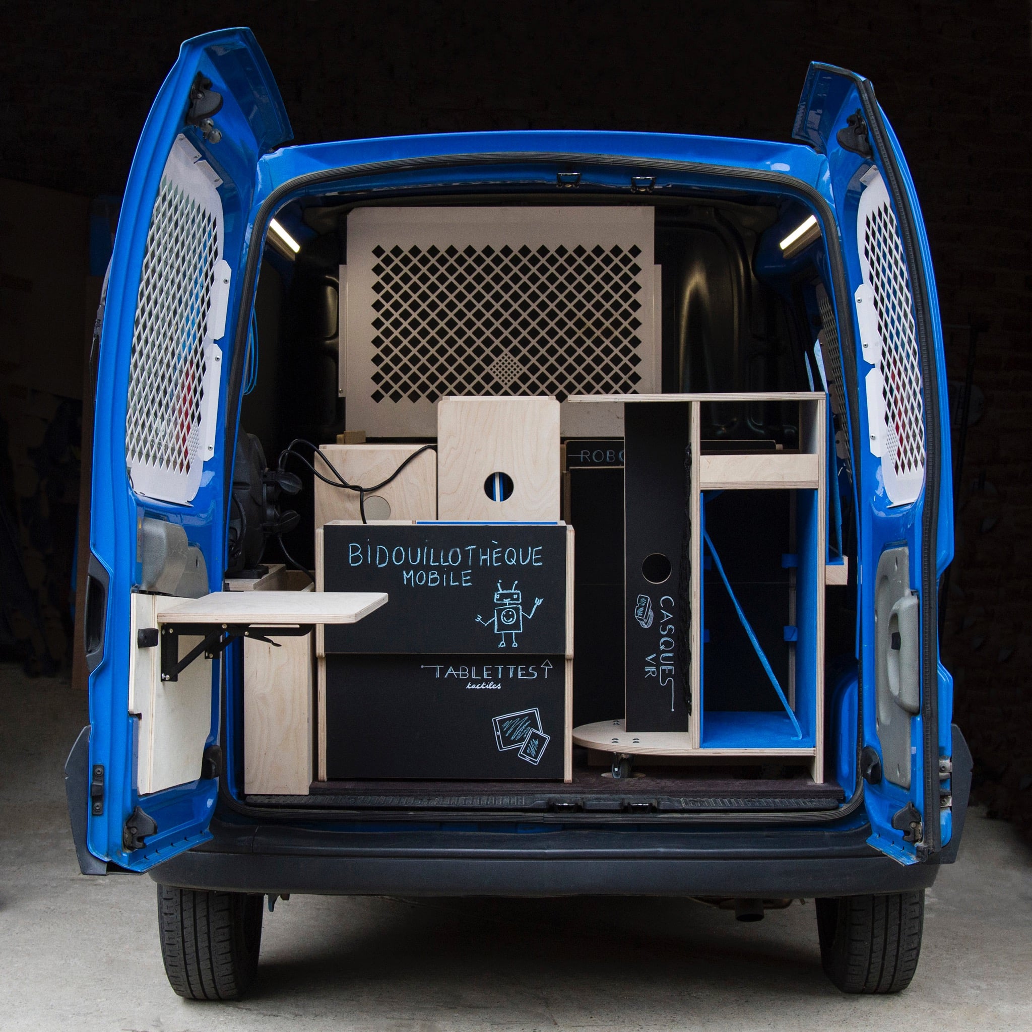 Vue de l'arrière du véhicule de la Bidouillothèque mobile, portes ouvertes, avec le mobilier en bois, bleu et noir.