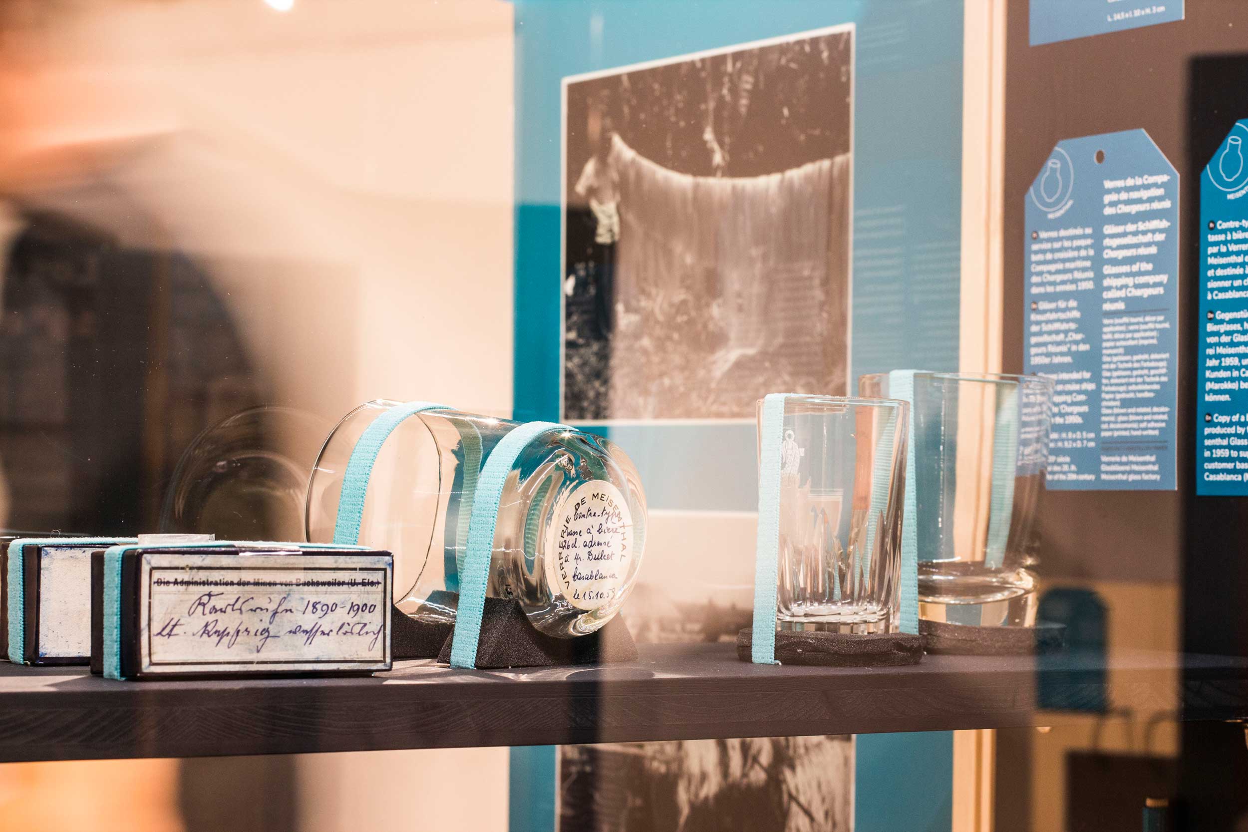 Gros plan sur des objets soclés en vitrine : une boîte d'échantillons de Bleu de Bouxwiller, et des verres de la Compagnie de navigation des Chargeurs réunis