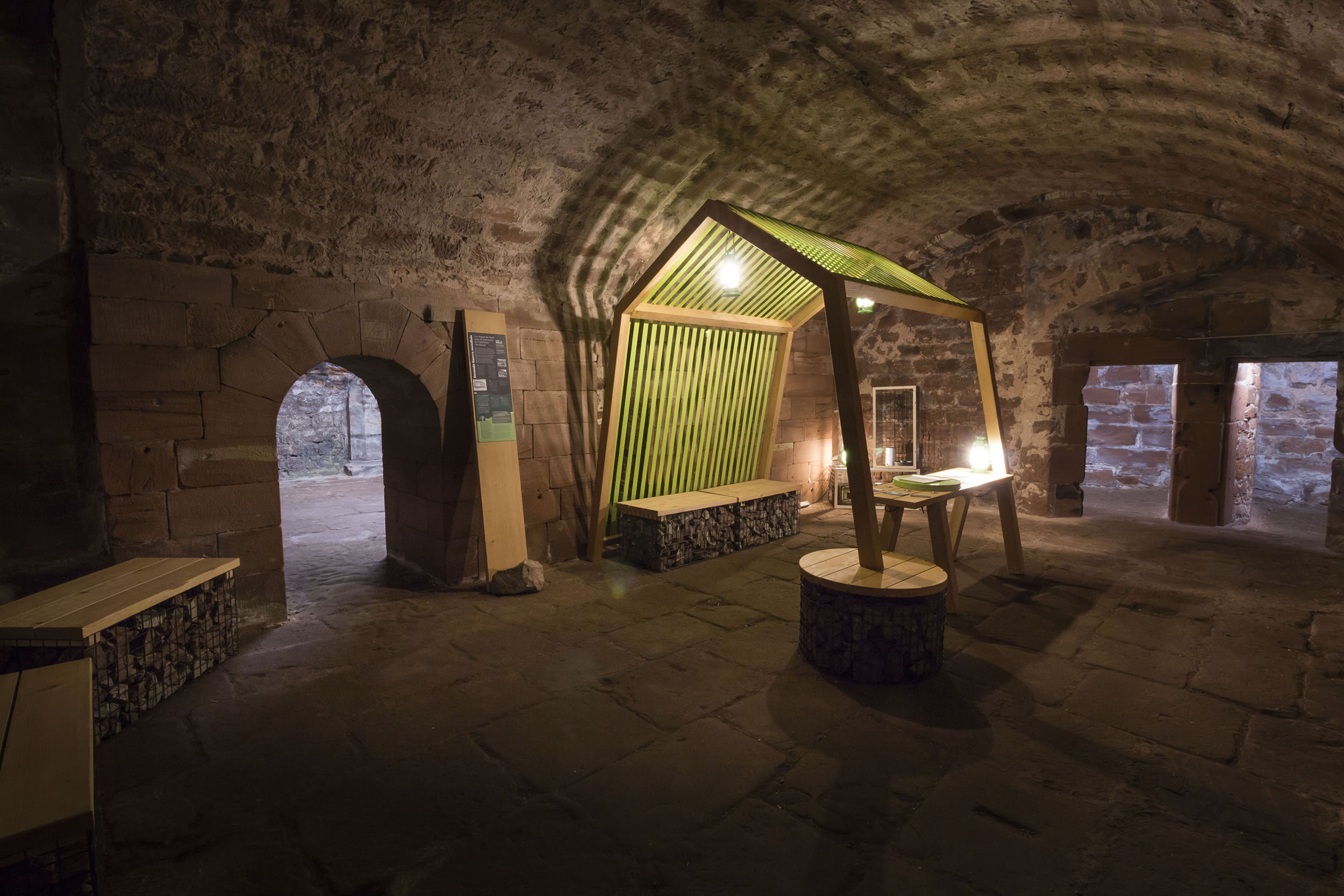 Dans une des casemates voûtées du château, vue d'ensemble d'une installation scénographique éclairée : une tente en bois et sangles vertes créé une sorte d'alcôve, sous laquelle les visiteurs peuvent lire des reproductions de documents historiques.