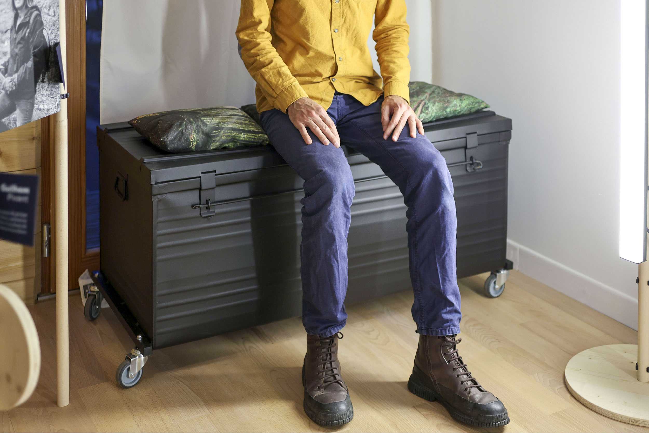 Un homme est assis sur une malle métallique noire, elle-même sur roulettes. Il doit s'agir d'une malle de rangement de la scénographie. Des coussins imprimés des photographies de la forêt, sont disposés sur la malle.