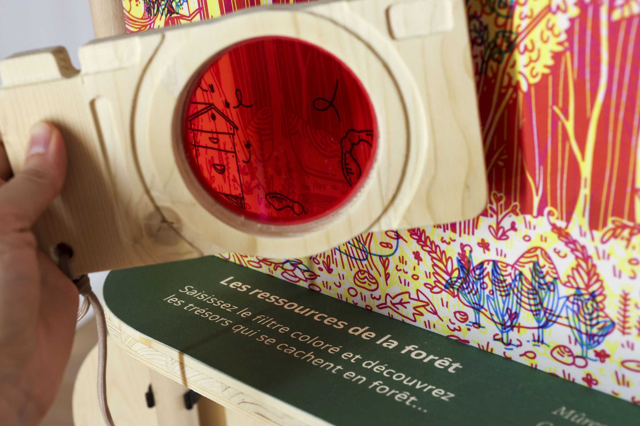 Un filtre rouge en forme d'appareil photo (le thème de l'exposition) est placé devant une illustration de "cherche et trouve". Des éléments sont cachés dans un paysage forestier. Le filtre permet de révéler les réponses.