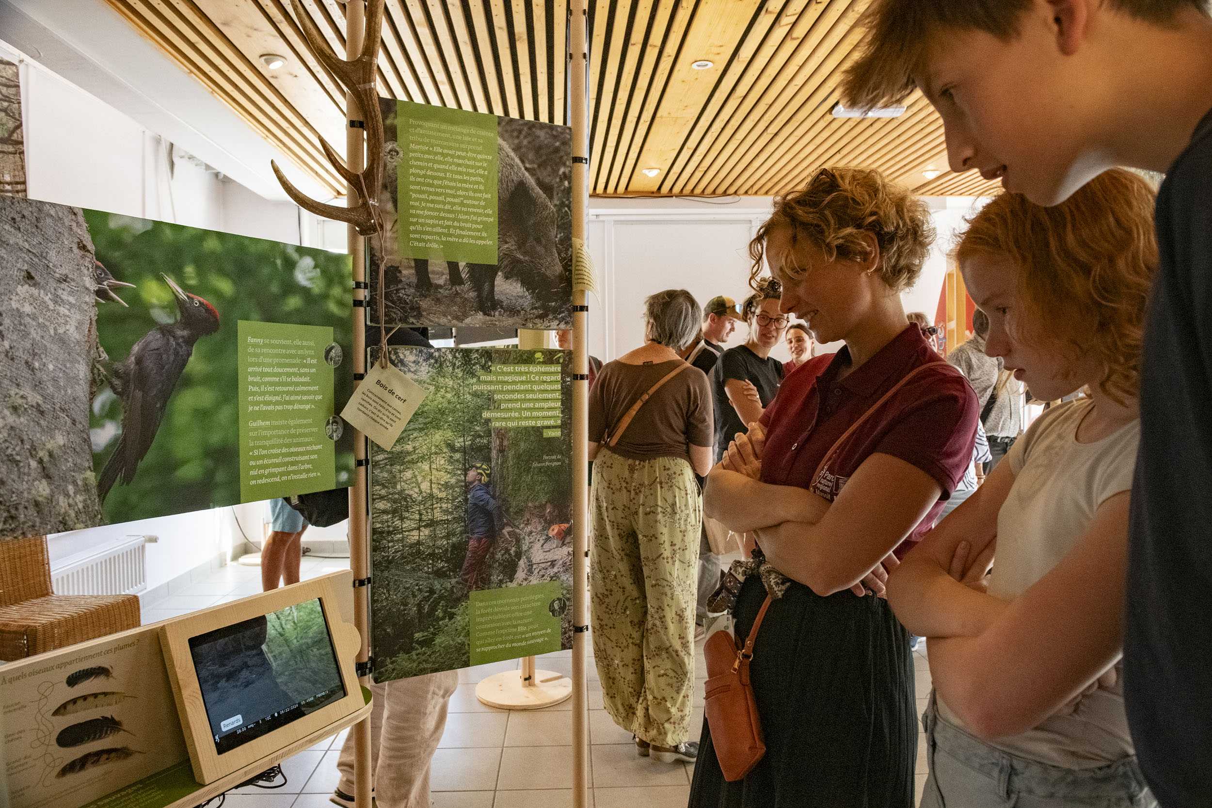 Trois personnes, une femme et deux adolescents, regardent attentivement une vidéo d'animaux filmés en pleine nature, diffusée sur la tablette. La tablette numérique est intégrée au mobilier scénographique, sous les tableaux de reproductions photographiques d'animaux et de personnages acteurs de la forêt.