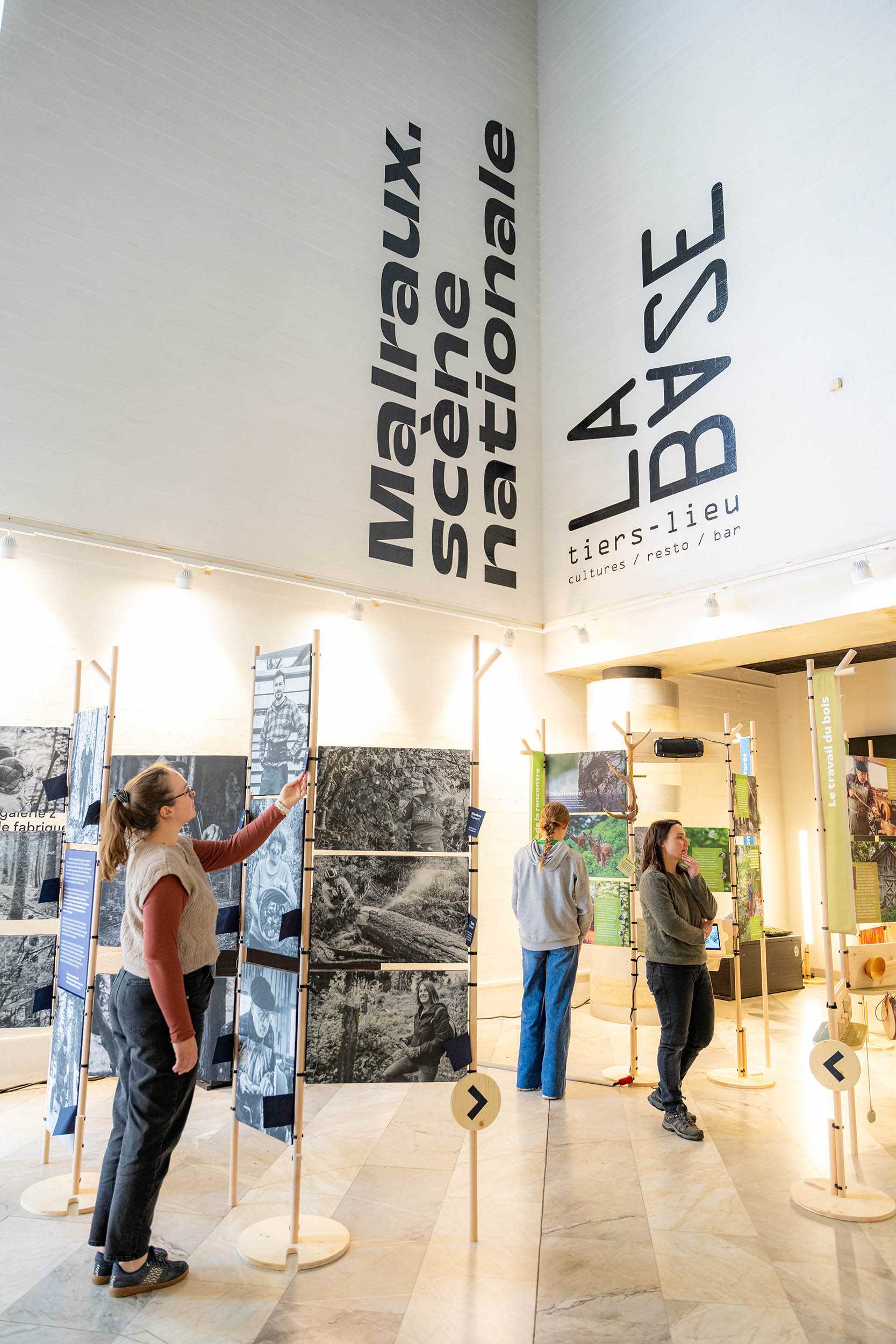Vue d'ensemble de l'exposition "Entre les branches, À l’affût des émotions partagées en forêt" dans la salle d'exposition du tiers-lieu "La Base", à la scène nationale Malraux de Chambéry. Les visiteurs posent en train d’observer les photographies reproduites en grand format.