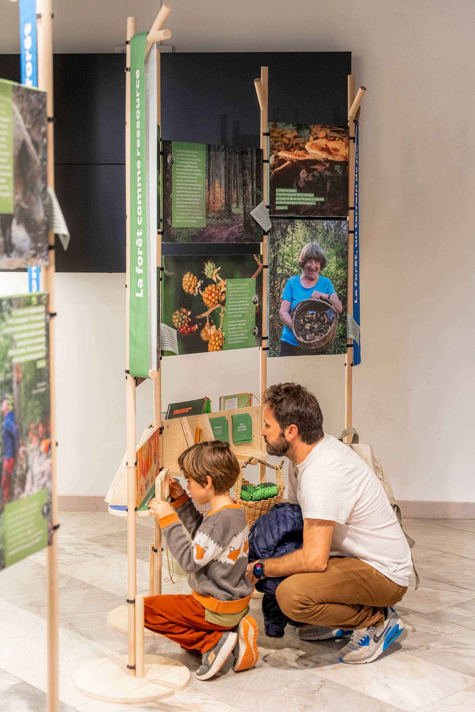 Un père et son fils sont accroupis au pied de l'exposition Entre les branches, conçue par le studio de design CYNARA, pour jouer à un jeu de cherche et trouve, à hauteur d'enfant.