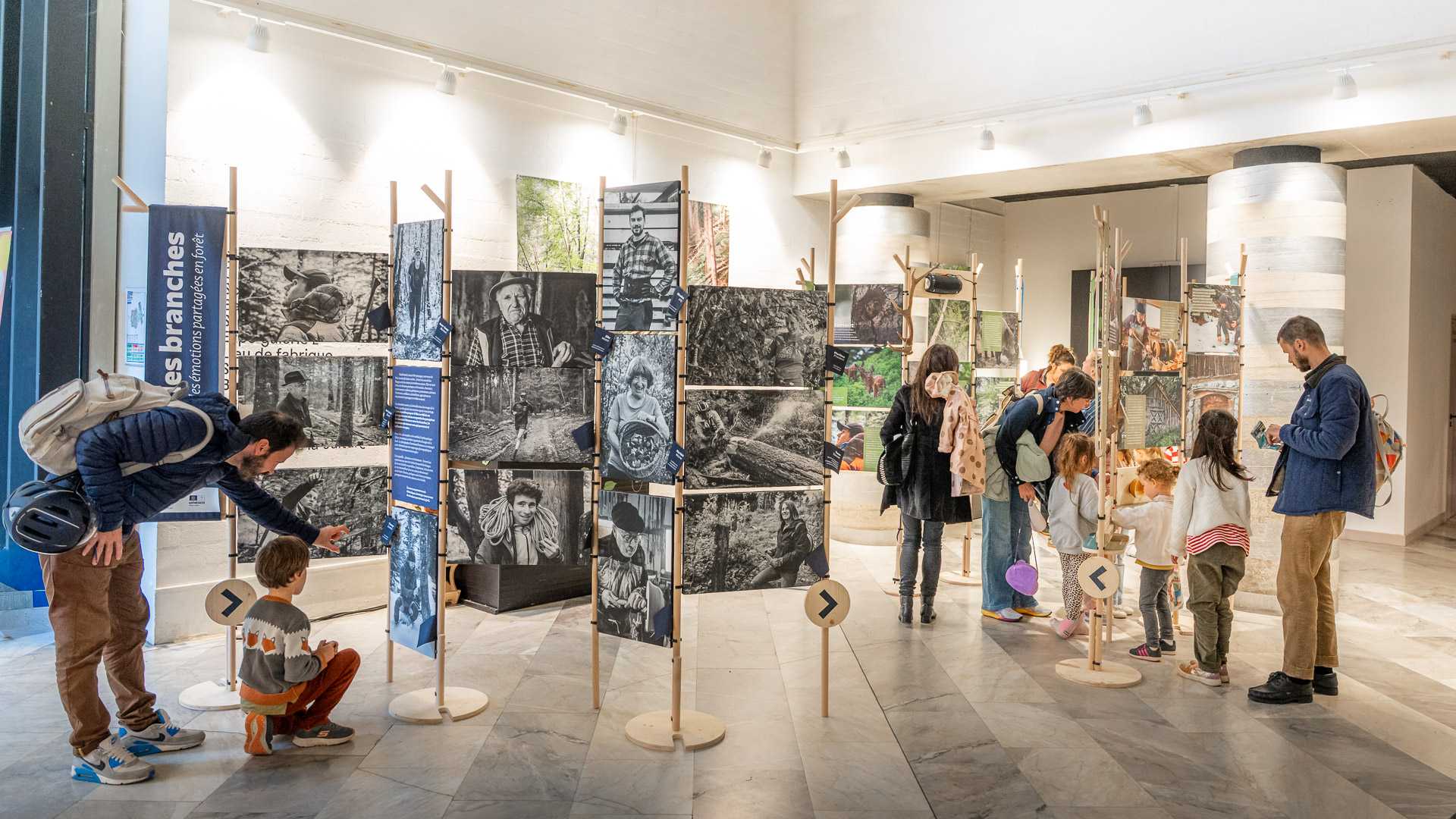 Vue d’ensemble panoramique de la scénographie d’exposition Entre les branches, avec les visiteurs venus en famille. Les parents lisent les cartels et les enfants font la queue pour jouer aux manipes.