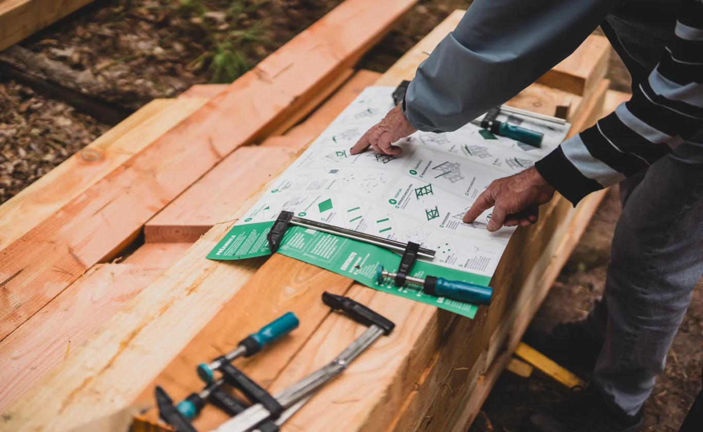 Vue rapprochée de la notice de construction, déployée, posée sur un tas de poutres en bois massif, avec des outils et des bénévoles qui montrent du doigt les étapes à suivre.