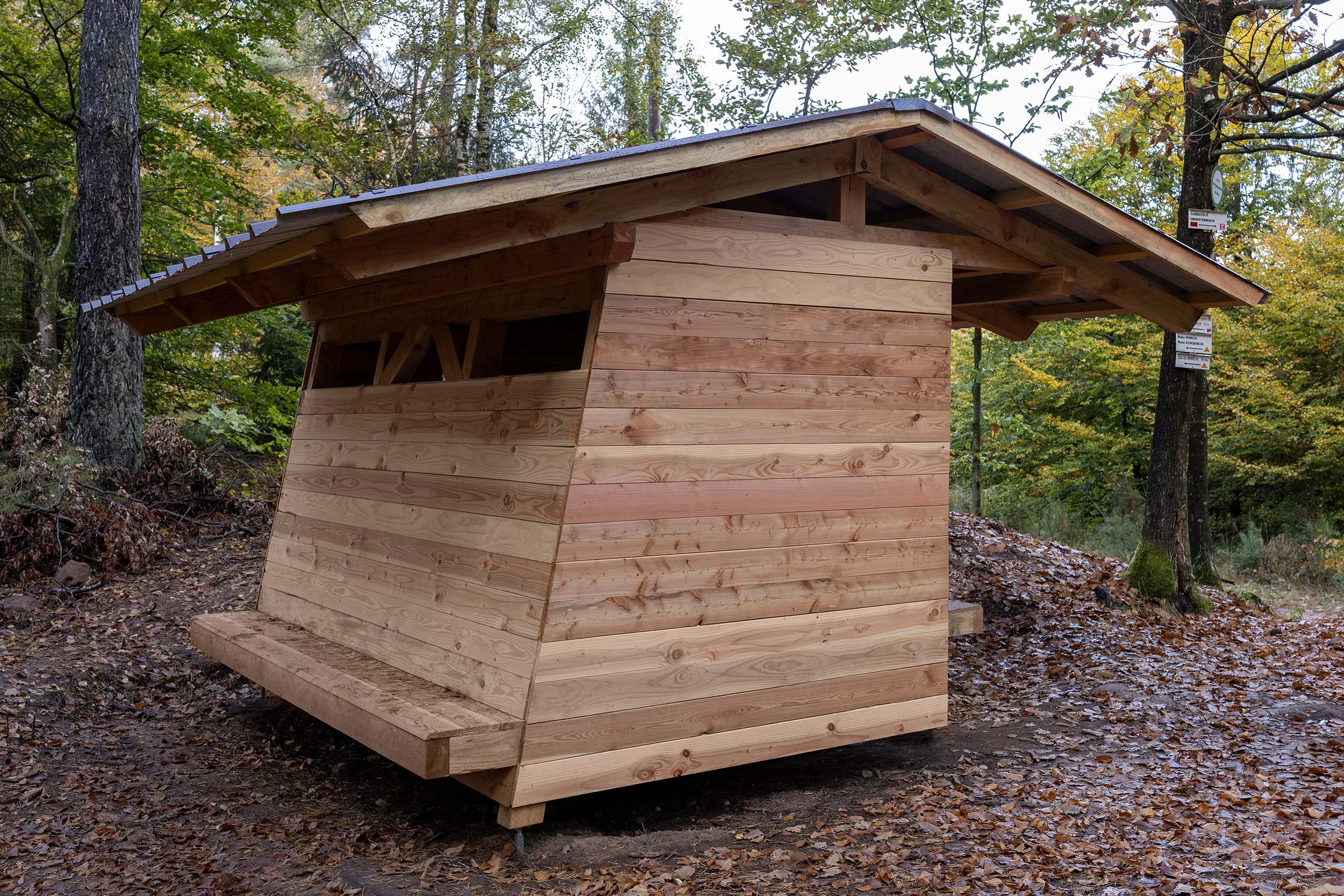 Vue de l'abri en entier, depuis son côté "parvis" sur le carrefour des chemins de randonnée. On voit la façade latérale avec le large banc incliné, protégé par le débord de toiture.