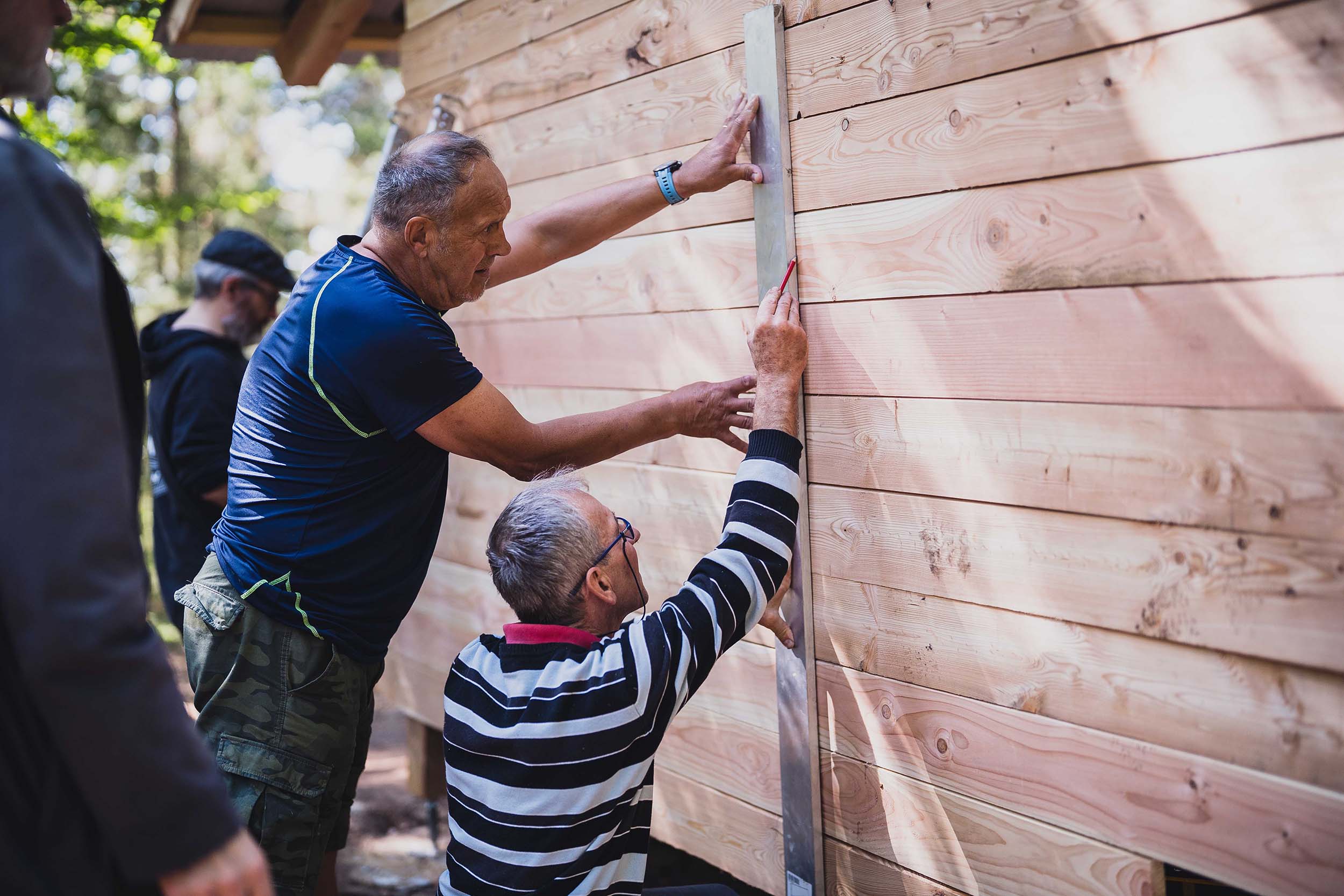 Deux bénévoles du Club Vosgien sont en train de tracer à la grande règle métallique sur le bardage extérieur, en vue de la création de l'ouverture pour la porte d'accès à l'abri.